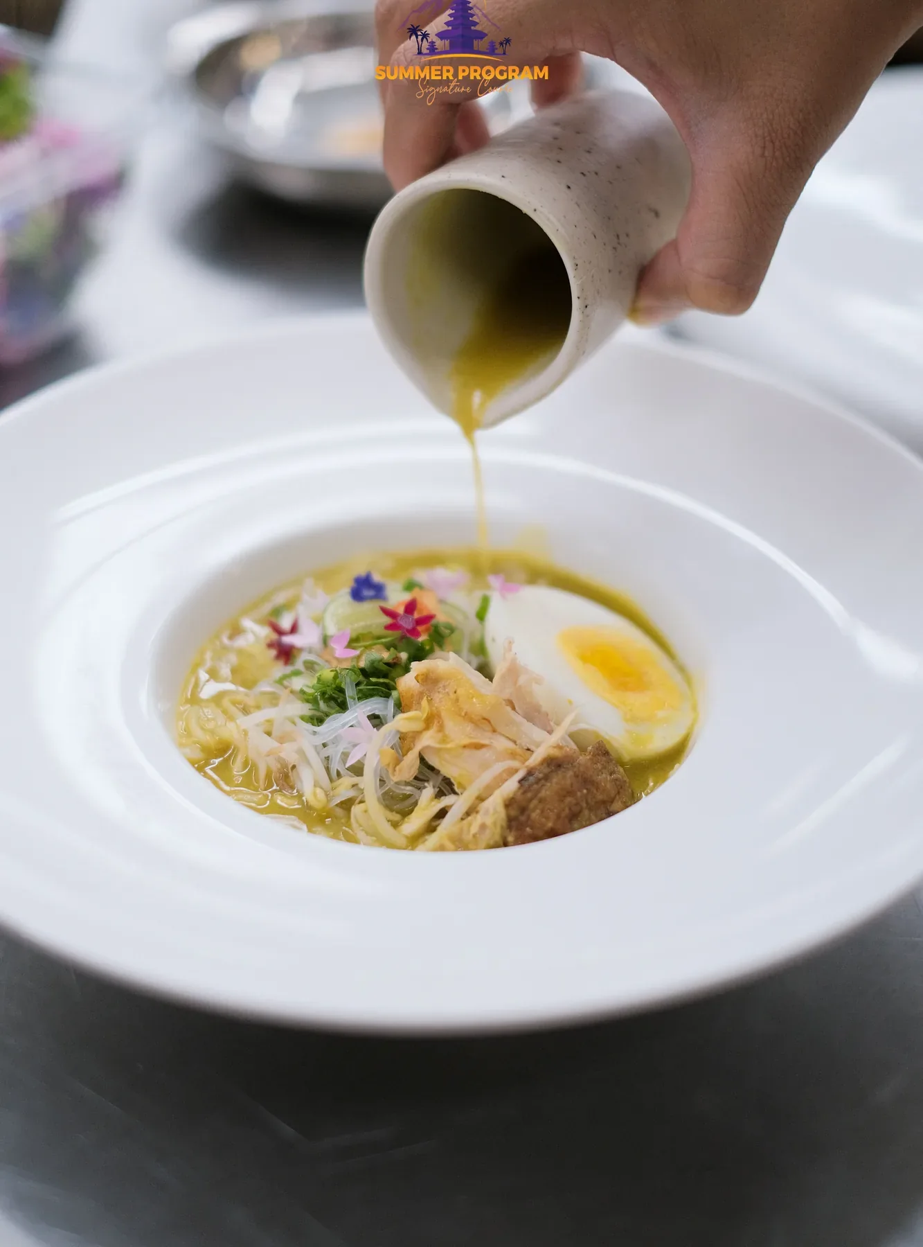 Hand pouring broth or sauce into a noodle and egg bowlic Culinary Arts program