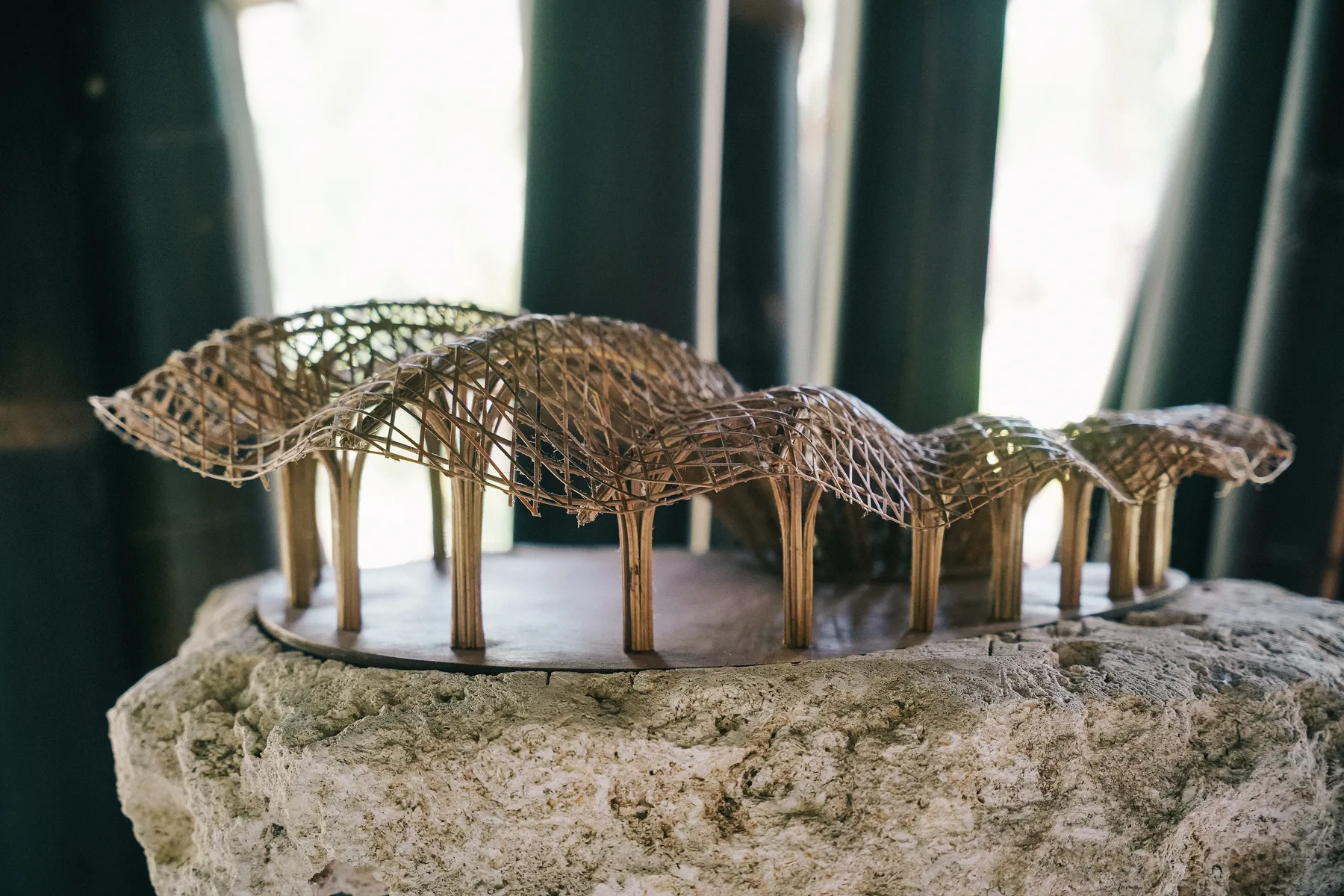 A detailed woven bamboo model of an undulating roof structure resting on a stone pedestal