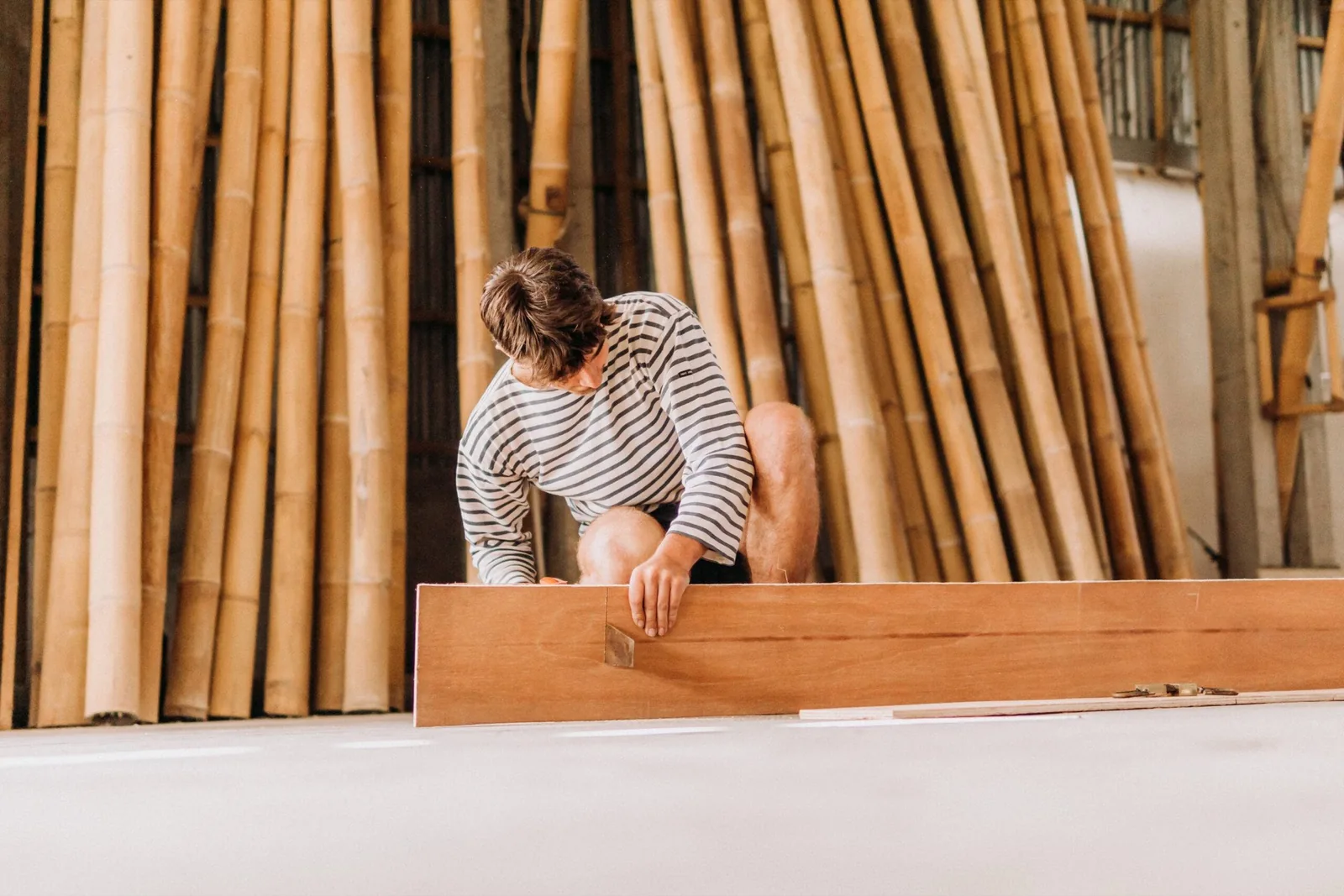 A student cutting timber in a Balinese workshop stacked with bamboo poles